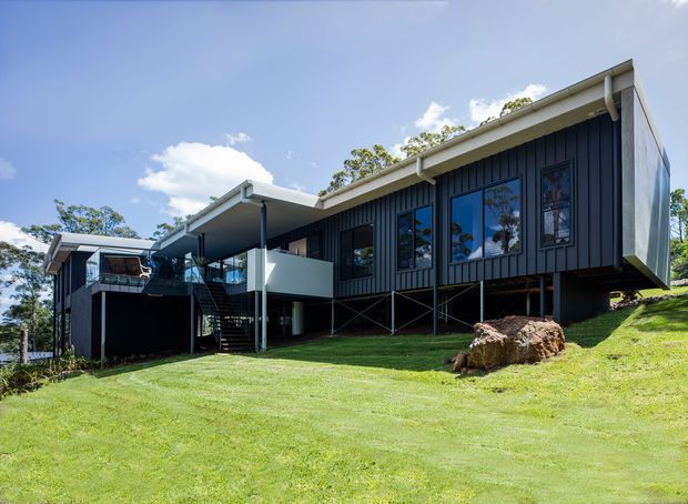 A contemporary dark-clad house set on a sloping grassy site, elevated on slender supports. The building has a strong horizontal form, mono-pitched rooflines, extensive dark-framed glazing, covered outdoor areas, and an exposed external stair connecting the levels. Bright daylight and mature vegetation surround the residence.