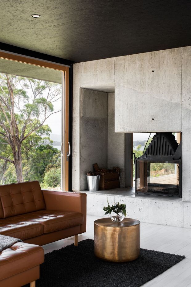 A contemporary living room with exposed concrete walls, ceiling, and fireplace surround. A large timber-framed window overlooks trees and a distant landscape. The room contains a brown leather sofa, black rug, cylindrical metallic coffee table, recessed ceiling light, and a built-in concrete fireplace with a black firebox.