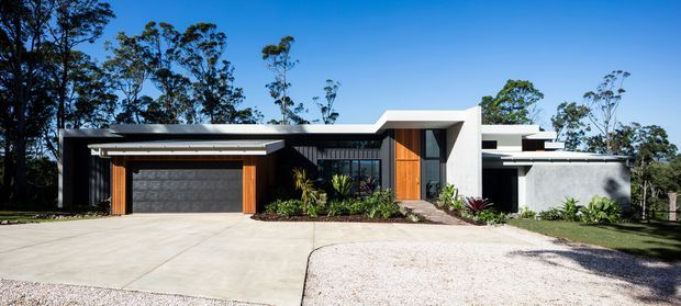 A contemporary single-storey residential facade set among trees, featuring a flat roof, dark vertical cladding, white rendered volumes, warm timber entry and garage detailing, and a broad pale concrete driveway with landscaped planting.