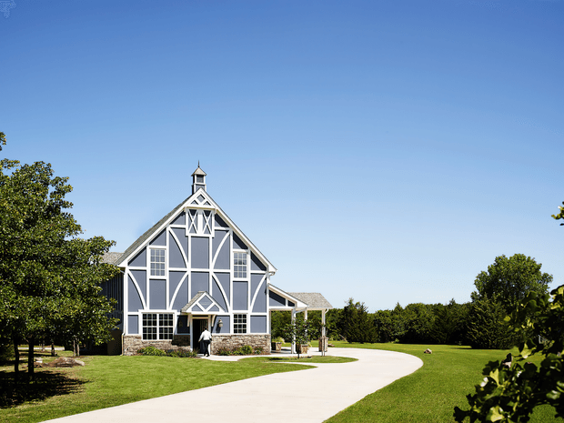 Exterior view of a large barn-inspired building with a steep gabled roof, prominent white timber-style framing over dark grey panels, tall multi-pane windows, and a small cupola. The building is set within expansive manicured lawns, mature trees, and a curved pale driveway under a clear blue sky.