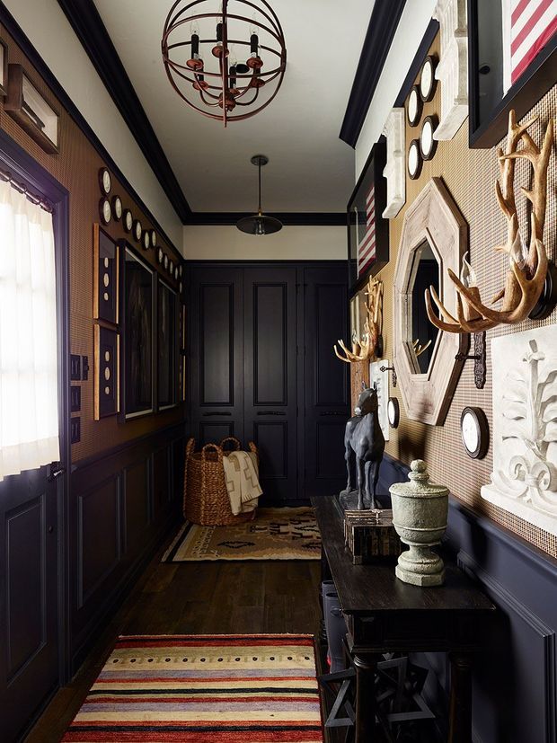 A narrow, richly detailed hallway or entry passage with dark navy-black raised-panel wall panelling and matching trim, patterned rugs over dark timber flooring, and a long console table. The walls feature framed decorative objects, clocks, sculptural animal figures, a large ornate wall feature, and woven-texture panels. A dramatic circular metal chandelier and smaller pendant light hang from the light ceiling. The space has a moody, eclectic, layered character with traditional architectural detailing.