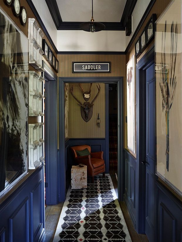 A richly detailed, narrow interior hallway with dark navy-blue painted wainscoting, door frames, and trim. The upper walls are covered in patterned wallpaper and lined with framed artwork and decorative objects. A sign reading “SADDLER” hangs above a doorway leading to a small seating nook with a leather armchair, patterned rug, and mounted antlers. The ceiling features dark ornamental trim and a central pendant light, creating a moody, historic, gallery-like atmosphere.