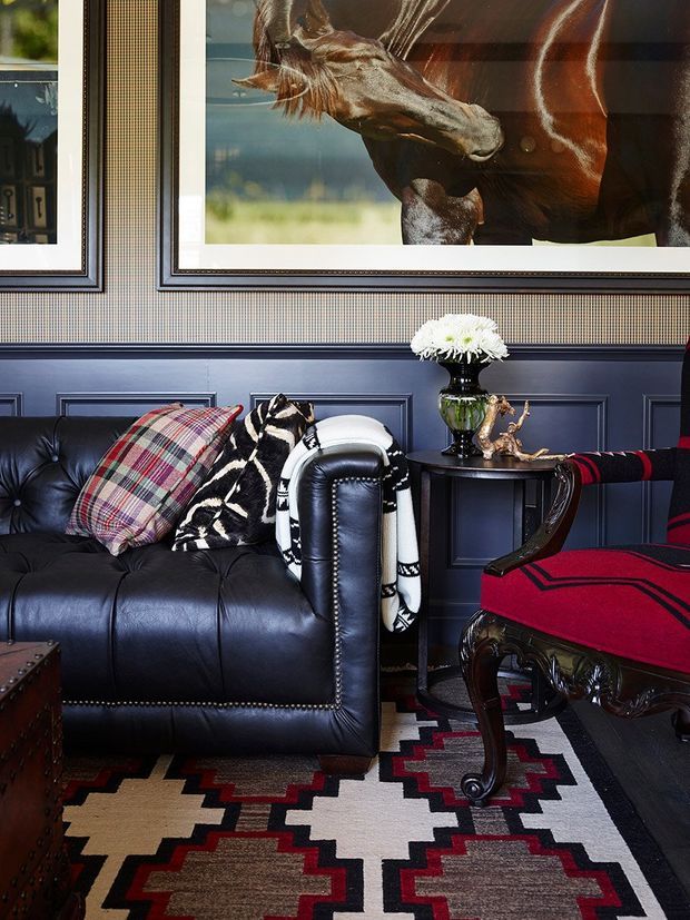 A richly styled living room with a black tufted leather sofa, a red upholstered traditional armchair, patterned cushions and throw, and a dark wood side table. The walls feature dark blue-grey decorative wainscoting beneath textured patterned wallpaper, with large framed animal artworks above. A bold geometric rug in black, cream, red, and brown anchors the room. The overall look is eclectic, dramatic, and traditionally inspired.