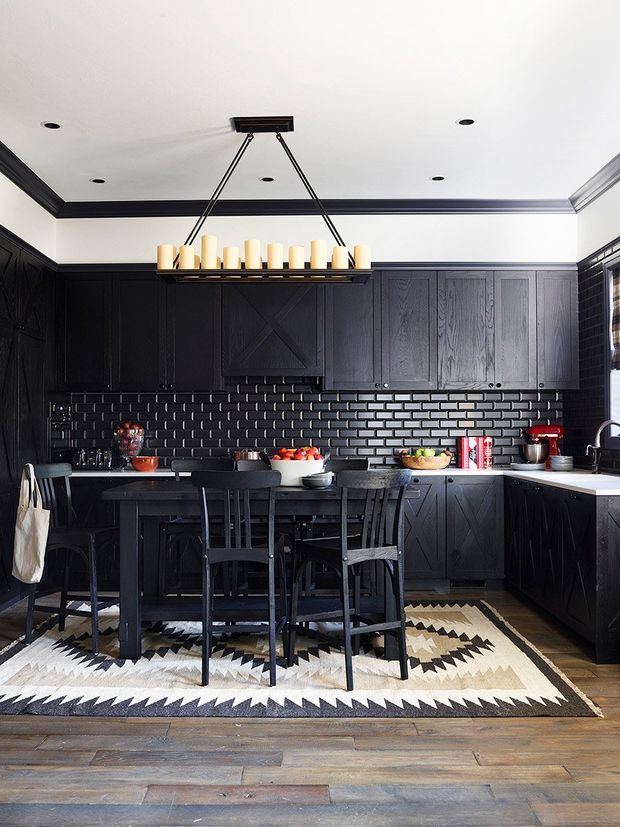 A dark, dramatic kitchen with black paneled cabinetry, a black subway-tile backsplash, pale benchtops, and a central dining island with black chairs. A linear pendant light hangs above the island, while a geometric black, cream, and tan rug contrasts with the timber floor. The room has a refined, moody traditional-meets-contemporary character.