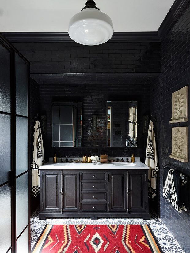 A dramatic, compact bathroom with black-painted brick walls, dark traditional-style double vanity cabinetry, twin basins, brass-toned tapware, framed mirrors, and a white globe pendant light. A bold red, black, and cream geometric rug covers the patterned floor, while monochrome patterned towels and decorative wall panels add visual interest. Frosted glass doors frame the space.