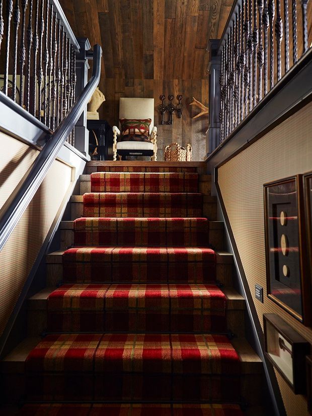 A richly detailed interior stairway viewed looking upward. Broad stairs are covered in red, rust, olive, and brown tartan carpet, bordered by dark timber treads and dark painted stringers. Ornate twisted balusters and dark handrails line both sides, with textured beige wall panels, framed artwork, and decorative objects at the upper landing. The landing has a dark timber plank floor and a traditional upholstered armchair, creating a warm, dramatic, heritage-inspired atmosphere.
