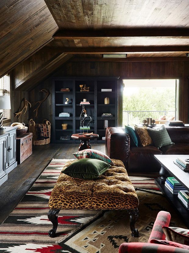 A richly layered living room with dark timber walls, floors, and an angled timber ceiling with exposed beams. A black built-in bookshelf displays books and objects beside a large window overlooking greenery. The room includes a deep brown leather sofa, patterned ottoman, colourful cushions, a patterned area rug, vintage-style cabinetry, and eclectic decorative objects. The overall atmosphere is warm, rustic, dramatic, and highly textured.