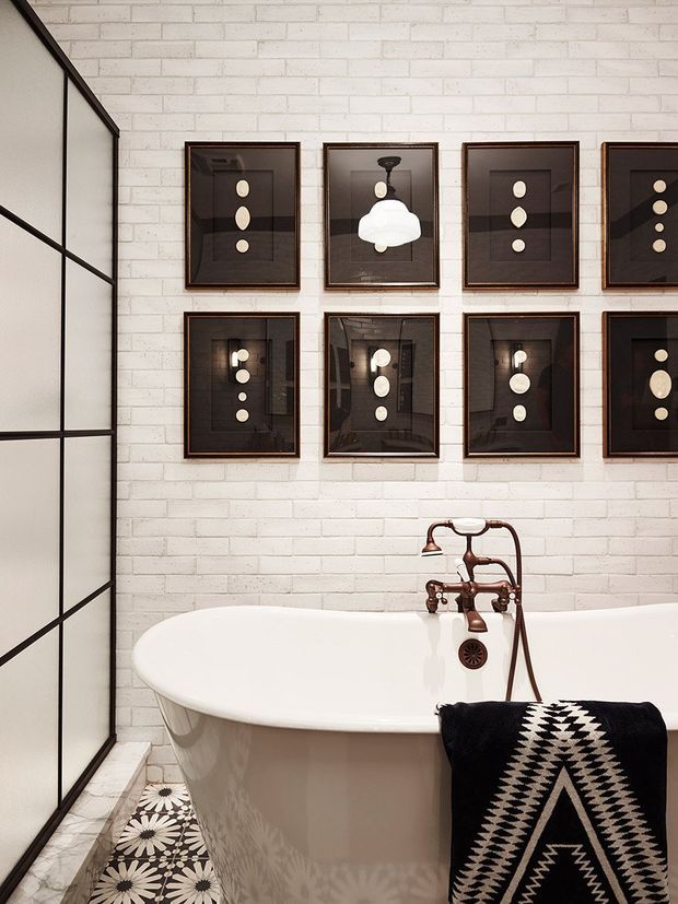 A stylish bathroom featuring a freestanding white clawfoot-style bathtub with an exposed copper-toned wall-mounted faucet and handheld shower. The room has white painted brick walls, black-framed translucent glazing or a shower screen at left, patterned black-and-white floor tiles, and a gallery arrangement of dark framed artwork above the tub. A black-and-white geometric towel adds contrast. The overall look is eclectic with industrial and vintage influences.