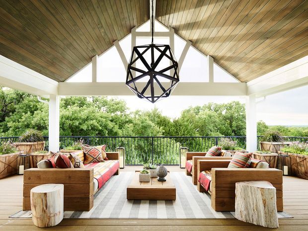 A covered outdoor lounge terrace with symmetrical timber-framed seating areas, colourful cushions, a striped area rug, a large black geometric pendant light, and a pitched timber-lined ceiling. White structural beams frame views of lush greenery beyond the black balustrade.