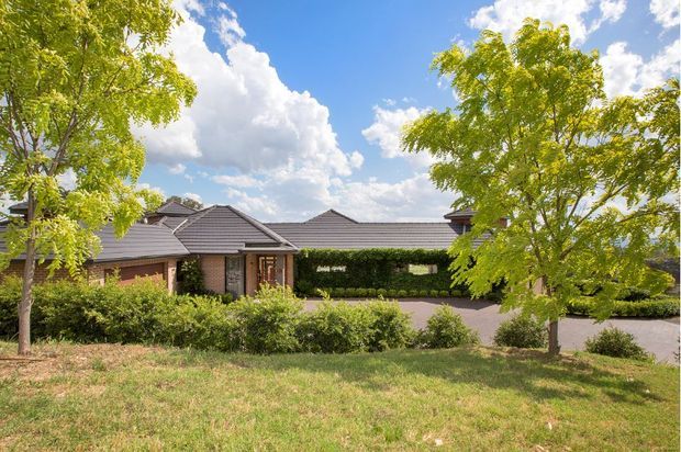 Single-storey residential house viewed from the street through landscaped front grounds. The home has a low, sprawling form with multiple hipped roof sections in dark grey tile, warm brick walling, stone-clad entry elements, covered parking or an entry canopy, and extensive mature planting. Bright daytime exterior photograph with blue sky and trees framing the facade.