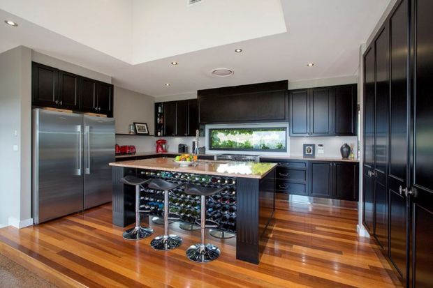A spacious contemporary kitchen with dark charcoal cabinetry, stainless-steel appliances, a polished stone island and integrated wine storage. The island includes seating for several people, while warm timber flooring, recessed lighting and a large horizontal window create a polished, inviting interior.