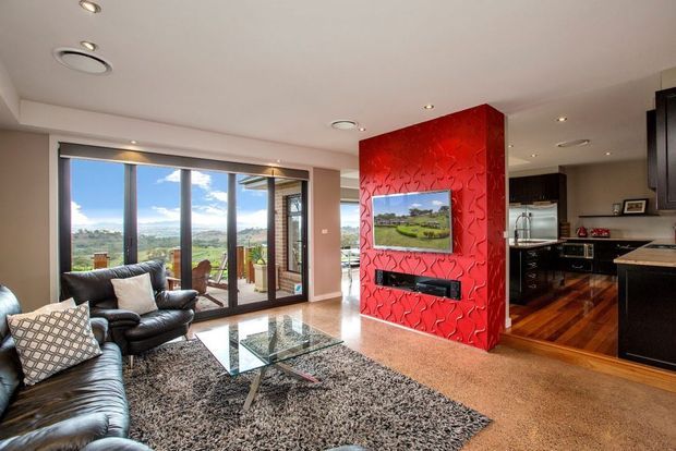Open-plan living room with black leather sofas, a glass coffee table, textured shag rug, and a striking red feature wall incorporating a mounted television and fireplace. Full-height glazed doors open to a scenic outdoor view, while a dark-toned kitchen with timber flooring is visible beyond the room.