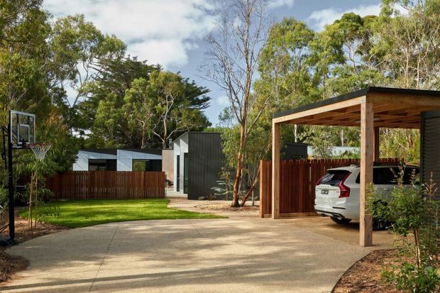 Exterior view of a contemporary residential property, showing a concrete driveway, timber-framed carport with a parked vehicle, dark corrugated wall cladding, timber fencing, landscaped lawn, and a basketball hoop. Mature trees surround the site, with a modern single-storey building visible beyond the driveway.
