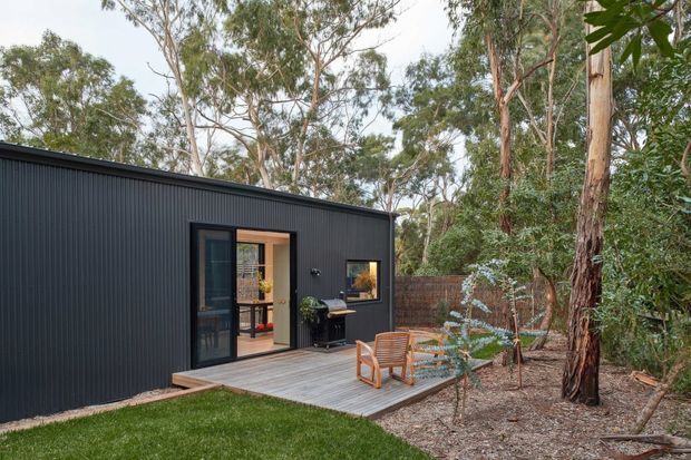 A compact single-storey modern dwelling clad in dark vertical ribbed metal sits within a eucalyptus woodland setting. The facade has a glazed dark-framed door and window opening onto a small timber deck with two outdoor chairs and a table. Native trees and a low brick boundary wall surround the home, creating a secluded natural outdoor area.