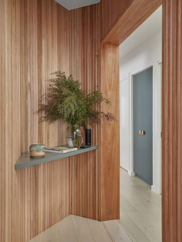 A refined residential entryway with floor-to-ceiling vertical timber wall panelling and matching timber-lined doorway. A slim muted green-grey floating shelf displays ceramics, books, a vase, and foliage. Through the opening is a bright white hallway with pale timber flooring and a grey-blue built-in door or cupboard with round timber handles. The space has a warm, natural, contemporary feel with strong linear detailing.