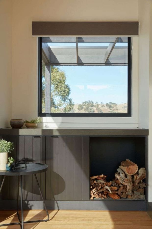 A compact, light-filled interior nook centred on a large black-framed window with a dark roller blind and a deep sill. Below the window is dark vertical-panel cabinetry with an open built-in compartment storing firewood. A small round side table with a potted plant sits in the foreground. The room has pale plaster walls, warm timber flooring, and views to a dry rural landscape outside.