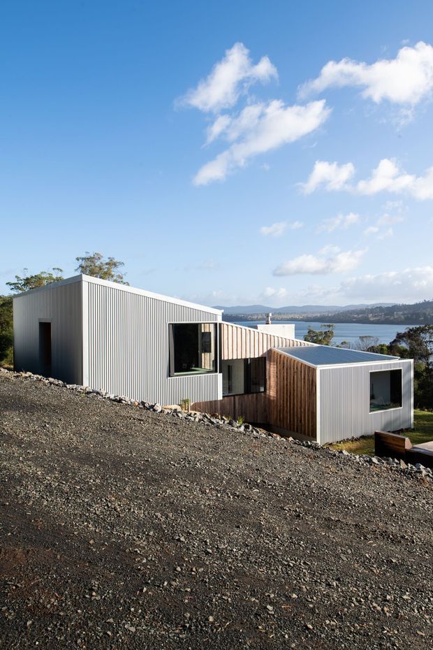 Exterior view of a modern, low-profile multi-volume home composed of corrugated metal-clad rectangular forms and a timber-clad central section. The building sits on a steep, rocky sloping site, with broad glazing and a distant body of water and hills visible behind it under a bright blue sky.