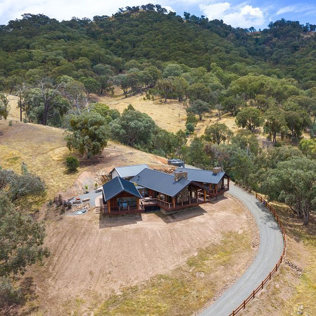 Aerial view of a substantial single-storey rural house nestled in a dry, wooded landscape. The building has dark pitched metal roofs, timber-framed walls and verandahs, multiple connected gabled volumes, and a curved gravel driveway with timber fencing. Dense eucalyptus-like bush covers the surrounding hills.
