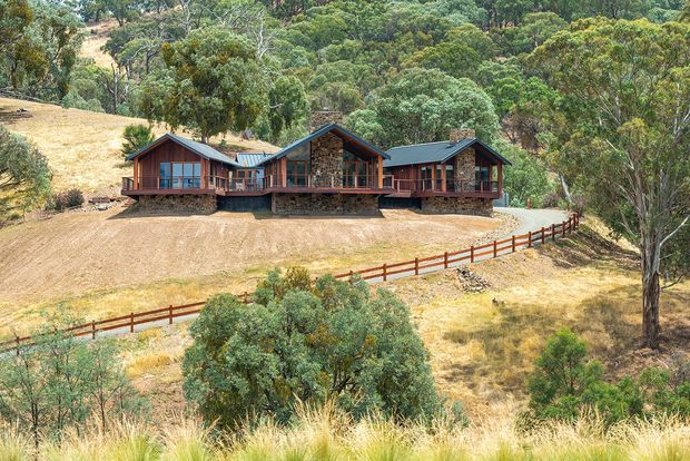 A wide exterior view of a rustic single-storey lodge-style home set on a dry, wooded rural site. The building has multiple connected wings, dark pitched gable roofs, extensive timber-framed glazing, deep verandahs, and prominent stone-clad wall sections. Timber fencing follows the sloping terrain in the foreground, with eucalyptus woodland surrounding the property.