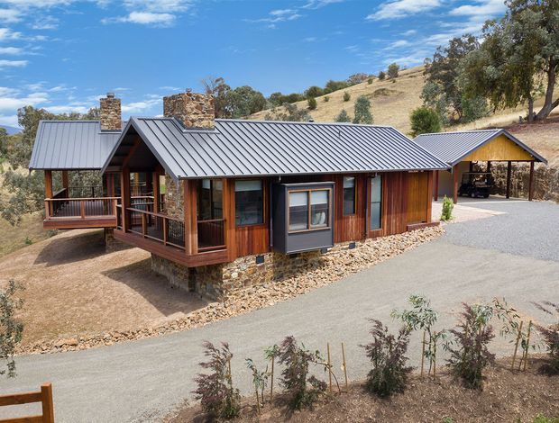 A single-storey timber-clad house set on a dry, sloping rural site. The building has a low-pitched corrugated metal roof, several stone-clad chimney structures, timber verandas and balustrades, and large areas of glazing. A detached or connected carport-like structure is visible at the right, with a gravel driveway in front. The landscape is sparsely planted with mature trees and dry grass under a bright blue sky.