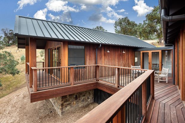 Exterior view of a timber-clad residential building arranged around an elevated deck and balcony walkway. The building has a long corrugated metal gable roof, vertical warm timber wall cladding, timber-framed glazing and doors, dark metal balustrades, and a stone masonry base. The surrounding dry, lightly wooded landscape and blue sky are visible beyond the structure.