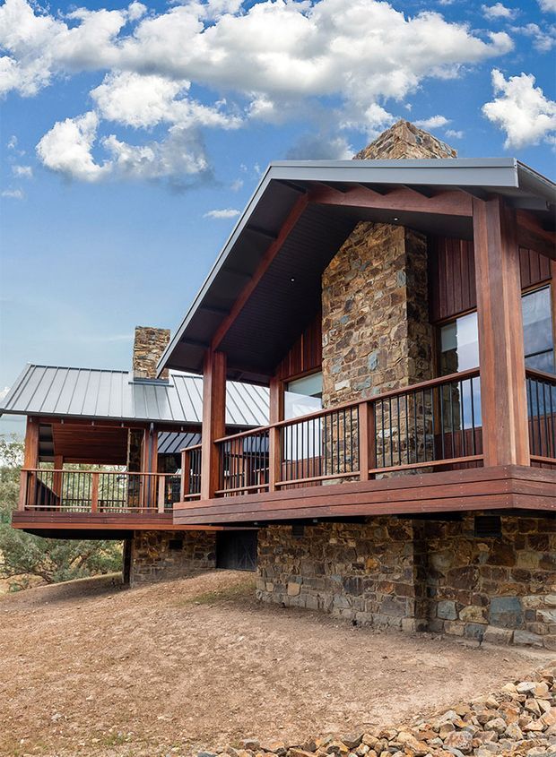 Exterior view of a substantial mountain-style house or cabin with elevated timber decks, dark timber posts and cladding, extensive rough natural-stone walls and chimney, and standing-seam metal pitched roofs. The building is set against a rocky mountainous landscape and blue sky.