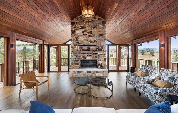 A spacious, warm living room with extensive floor-to-ceiling glazing and timber framing, opening to views of a dry rural landscape. The room features a full-height rough-stone fireplace chimney with a black fireplace insert, a richly timber-lined sloping ceiling, timber flooring, floral upholstered sofas, a leather lounge chair, and round glass-and-metal coffee tables. A sculptural pendant hangs above the fireplace, creating a rustic yet refined retreat-like atmosphere.