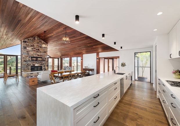 A spacious open-plan kitchen overlooking an adjoining dining and living area. The room features a large white island with shaker-style drawers, white perimeter cabinetry, dark timber flooring, and a striking warm timber-plank ceiling that curves around a substantial exposed stone fireplace. Extensive glazing and timber-framed doors bring in natural light and views of the surrounding landscape. Pendant lights hang above the dining area, while recessed black downlights illuminate the kitchen.