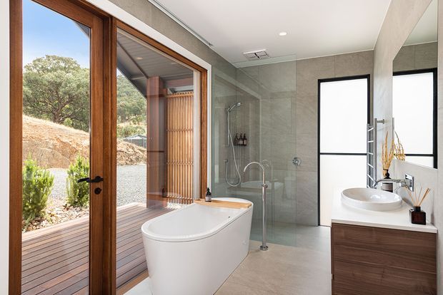 Contemporary bathroom with a freestanding white bathtub, glass-enclosed shower, wall-mounted round basin, and dark timber vanity. Large timber-framed glazed doors open to a small exterior deck and landscaped setting, while pale stone-look wall and floor finishes create a clean, light-filled interior.