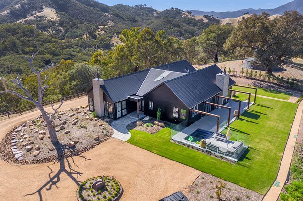 Aerial view of a contemporary dark-clad single-storey residence set within a landscaped mountain or bush setting. The home has multiple pitched roof volumes, extensive glazing, a covered outdoor entertaining area, manicured lawns, paved paths, gravel garden areas, and a small rectangular pool or water feature beside the house.