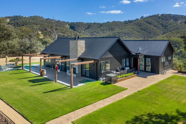 A single-storey dark-clad house set within a landscaped rural or bush setting. The building has multiple pitched gable roof forms with dark metal roofing, large glazed openings, a central stone-clad chimney or feature wall, and an outdoor covered entertaining structure with timber posts. Broad manicured lawns and gravel paths surround the home, with wooded hills visible behind it.