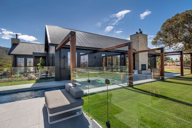 Exterior view of a contemporary single-storey residence with a steep pitched corrugated metal roof, dark vertical cladding, warm timber structural beams, extensive glazing, and a prominent stone chimney. The landscaped outdoor area includes a tiled terrace, glass balustrade, manicured lawn, covered timber-framed outdoor zone, and outdoor seating. Mountainous terrain is visible in the distance.