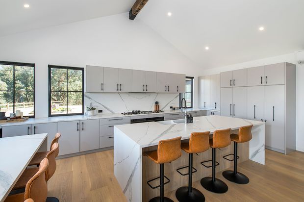 Bright, spacious contemporary kitchen with a large white marble-look island, four upholstered bar stools, extensive pale cabinetry, integrated appliances, black-framed windows, timber flooring, and a partially exposed timber ceiling beam. A dining area is visible at the left edge.