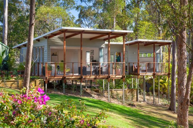 Exterior view of a low-set, elevated residential building composed of connected lightweight pavilion-like sections in a leafy landscaped setting. The home features pale horizontal cladding, broad shallow roof overhangs, exposed timber posts and beams, and raised timber verandahs with slim metal balustrades. Dense trees and garden planting surround the structure.