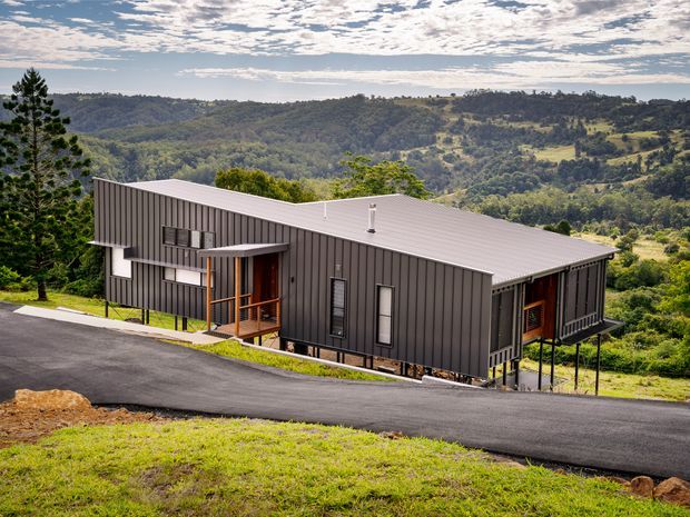 A contemporary single-storey dwelling set on a steep, elevated rural hillside. The elongated building has dark vertical metal cladding, a low-pitched roof, extensive glazing, and raised sections supported on slender posts. A paved driveway curves across the foreground, with expansive wooded and pastoral hills beyond.