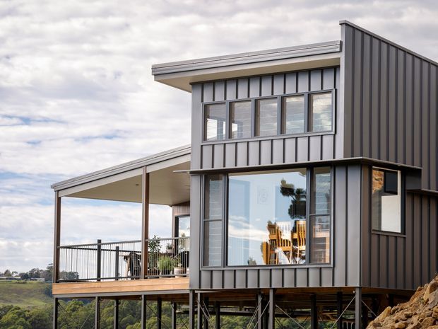 Exterior view of a contemporary multi-level house on a sloped site, elevated on steel supports. The building has dark vertical metal cladding, extensive black-framed glazing, a projecting upper volume, and a covered outdoor deck with slim metal railings. A mono-pitched roofline and angular asymmetric form are prominent, with rural landscape visible beyond.