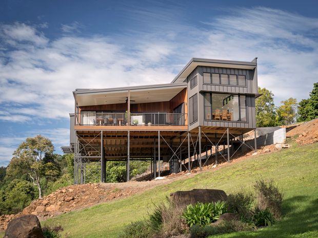 Exterior view of a contemporary multi-level house elevated on slender supports above a steeply sloping, landscaped site. The building has dark vertical cladding, extensive glazing, a broad skillion-style roof, and a large covered balcony with timber decking and metal balustrades. The exposed undercroft structure and hillside setting are prominent.