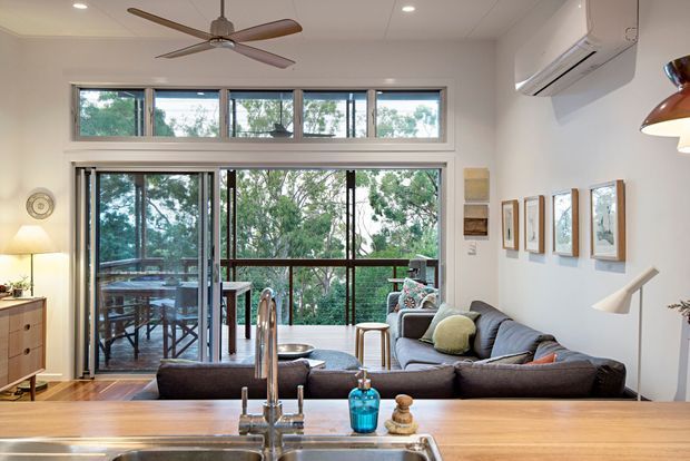 Open-plan living area viewed from a kitchen, with a timber benchtop and sink in the foreground. The room features a large grey sectional sofa, sliding glass doors opening to a timber outdoor deck, clerestory windows, ceiling fan, split-system air conditioner, and framed artwork. The interior is bright and connected to a leafy outdoor setting.