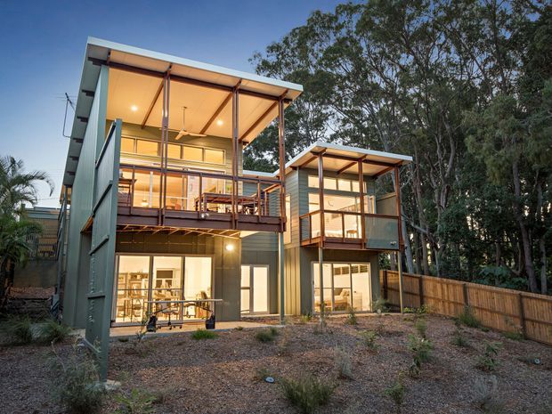 Exterior view of a contemporary multi-level residential building with two connected or closely positioned wings, elevated timber balconies, extensive glazing, exposed slender posts and beams, and asymmetrical skillion-style roof forms. The structure is set among established trees with a timber boundary fence and landscaped gravel foreground. Warm interior lighting is visible through the windows and the upper-level covered outdoor areas.
