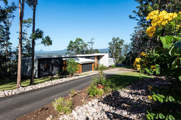 Exterior view of a contemporary single-storey home set within a landscaped, sloping site. The building has dark vertical cladding, a light flat-roofed projecting section, broad glazing, and a paved driveway approached through native planting and rock beds. A distant mountain landscape is visible beyond the property.