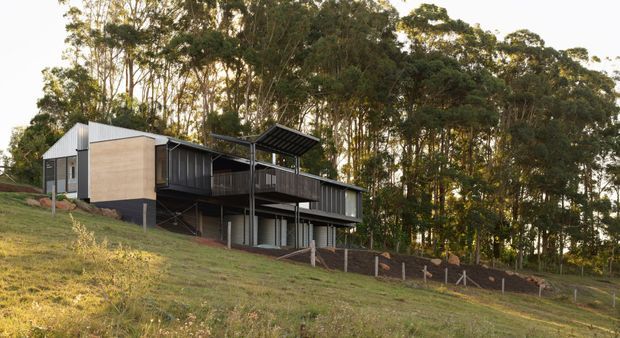 A modern multi-level house set into a sloping, wooded site. The building features dark vertical cladding and glazing, a light rendered or concrete upper volume, projecting decks and balconies, slender structural posts, and a pitched metal roof with a prominent covered outdoor structure. The surrounding eucalyptus-like woodland and elevated hillside setting are clearly visible.