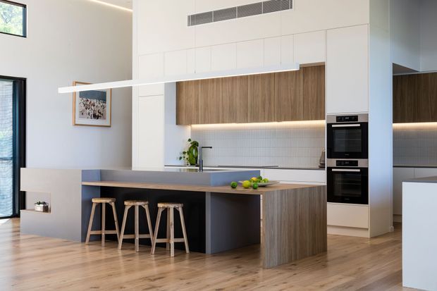 A bright, contemporary open-plan kitchen with a large island and integrated seating for four. The kitchen features flat-panel white and timber-look cabinetry, a dark island base, a pale stone benchtop, a small light-grey tiled splashback, integrated double ovens, timber flooring, and linear pendant lighting. Tall glazing and high ceilings contribute to the airy feel.