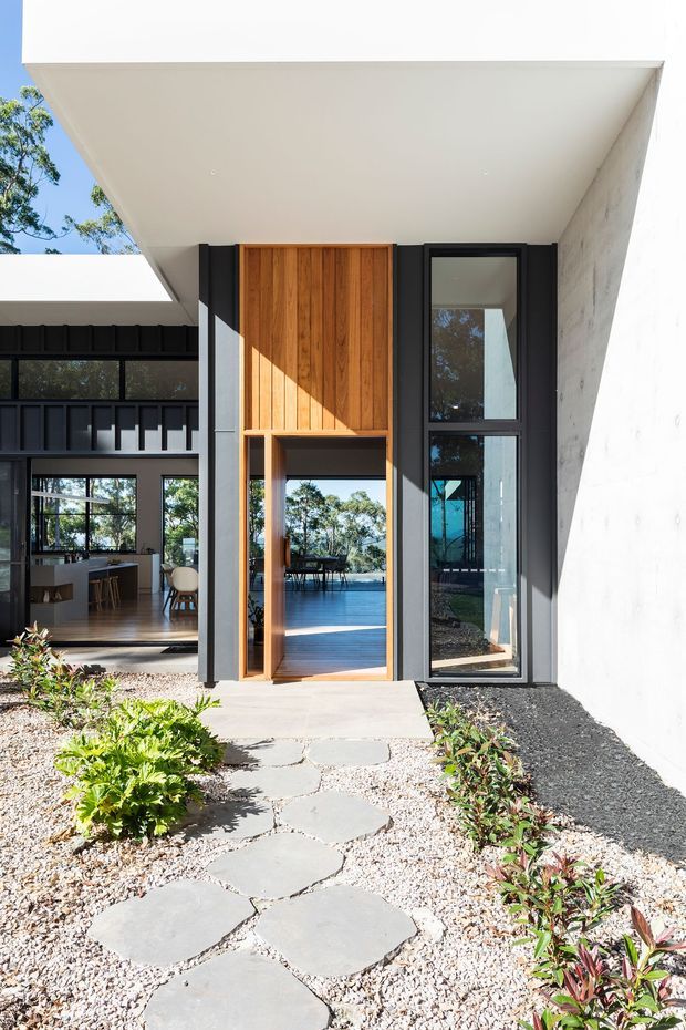 A bright contemporary residential exterior viewed from a landscaped front path. The entry features a tall vertical timber door and timber-clad panel set within dark-framed glazing, beneath a dramatic projecting white concrete or plastered roof volume. An open-plan interior with kitchen and dining furnishings is visible through the adjacent glazing, while stepping stones, gravel, and low planting lead to the entrance.