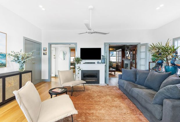 Bright, airy living room with white walls and ceiling, pale blue-grey trim, a central fireplace with wall-mounted television, large blue sectional sofa, cream lounge chairs, and a warm rust-toned area rug. Open doorways connect to adjacent rooms, while a ceiling fan, plants, framed artwork, and decorative cabinetry add detail. The room has a relaxed coastal-contemporary feel with strong natural light.