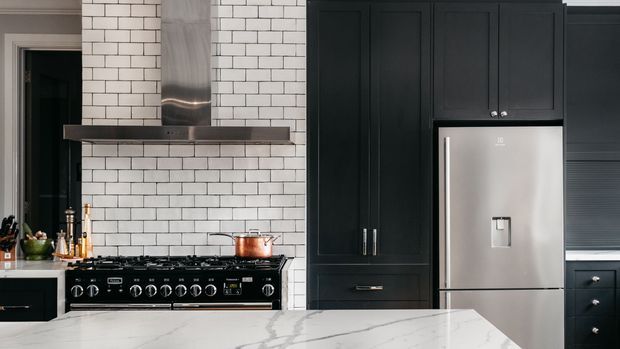 A refined kitchen with a large white marble-look island in the foreground, black shaker-style cabinetry, a stainless-steel range and refrigerator, and a white subway-tile backsplash. Brass-toned hardware and cookware add subtle warmth to the predominantly monochrome scheme.