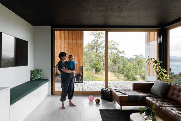 A contemporary living room with a dark textured ceiling, pale timber flooring, white built-in cabinetry and a green upholstered bench beneath a wall-mounted television. Large timber-framed glazed doors open onto a timber deck and overlook trees and a body of water. A brown leather sectional sofa, indoor plants and children's toys create a relaxed family setting.