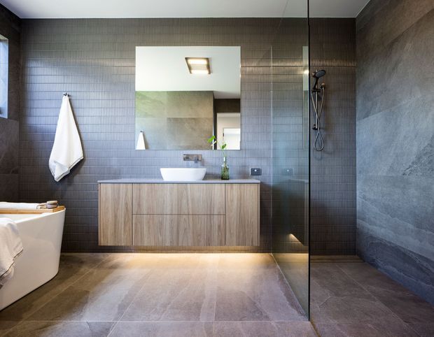 Contemporary bathroom with a freestanding white bathtub, floating timber vanity, wall-mounted basin, large mirror, walk-in glass shower, and dark grey tiled walls. The space has a refined, neutral palette with warm timber joinery and concealed under-vanity lighting.