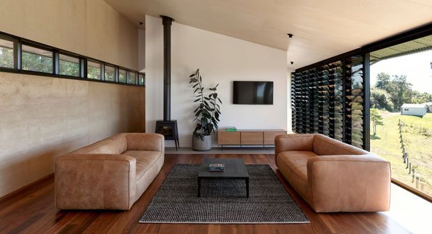 A warm, contemporary living room with two tan leather sofas facing each other across a dark coffee table and textured rug. The room features rich timber flooring, a plastered feature wall with high-level clerestory windows, expansive black-framed glazing with external louvres, a wall-mounted television, low timber media cabinetry, a freestanding wood-burning stove, and a potted plant. The glazing opens to a rural landscape, creating a bright indoor-outdoor connection.