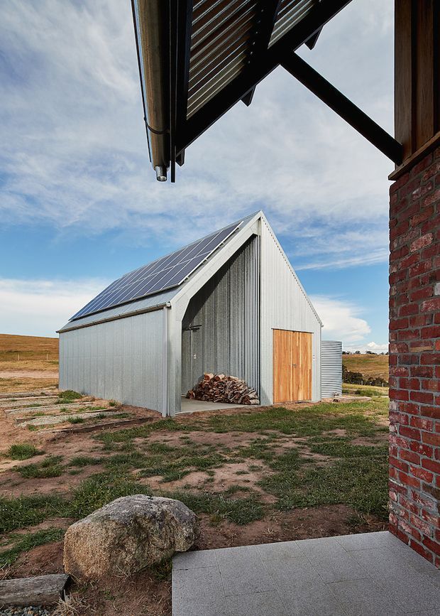 Exterior view of a small corrugated-metal gable-roof outbuilding with a large array of solar panels on one roof slope. The structure has a ribbed grey metal facade, a timber double door, and an open storage bay containing firewood. A brick building edge, covered roof structure, tiled paving, rural paddocks, and open sky surround the shed.