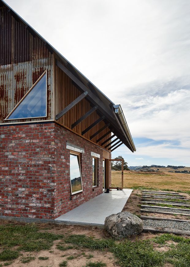 Exterior view of a rural, barn-like building with weathered red brick walls, corrugated metal and dark timber cladding, a steep gabled roof with exposed timber framing, and a distinctive triangular window. A covered veranda with concrete paving runs along the side, opening toward expansive dry farmland and low hills.